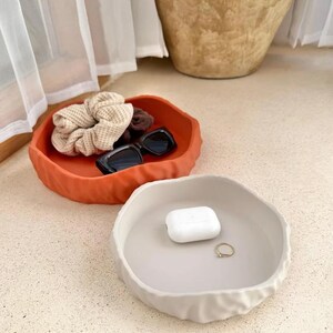 May include: Two round, shallow trays with wavy, textured edges. One tray is a vibrant terracotta orange, holding a beige waffle-knit scrunchie, a dark brown scrunchie, and black sunglasses. The other tray is a neutral cream, holding white earbuds.