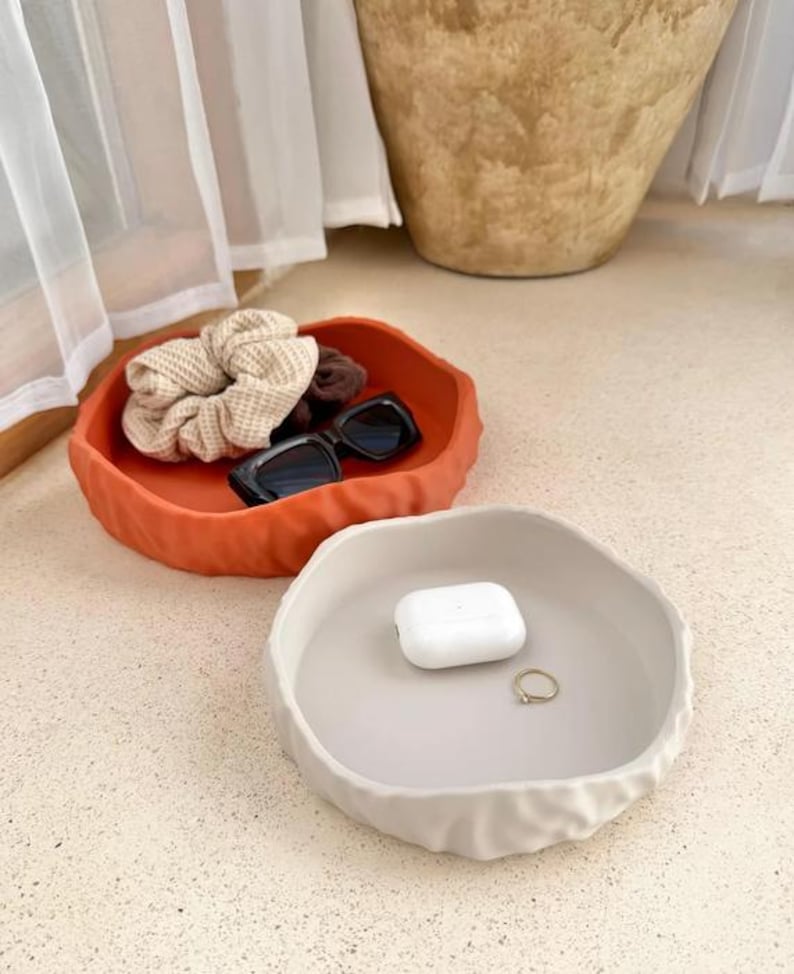 May include: Two round, shallow trays with wavy, textured edges. One tray is a vibrant terracotta orange, holding a beige waffle-knit scrunchie, a dark brown scrunchie, and black sunglasses. The other tray is a neutral cream, holding white earbuds.