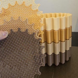 May include: A multi-layered, decorative planter with a wavy design. The planter is composed of alternating bands of beige, gold, and brown. A matching, openwork, star-shaped piece is held in front, showcasing the intricate pattern.