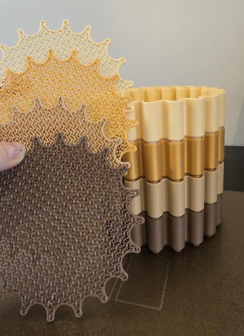 May include: A multi-layered, decorative planter with a wavy design. The planter is composed of alternating bands of beige, gold, and brown. A matching, openwork, star-shaped piece is held in front, showcasing the intricate pattern.