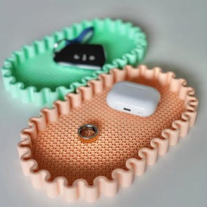 May include: Two decorative trays with a wavy edge. One is light green, holding keys. The other is peach-colored, holding a ring and a white wireless earbud case. The trays have a textured surface.