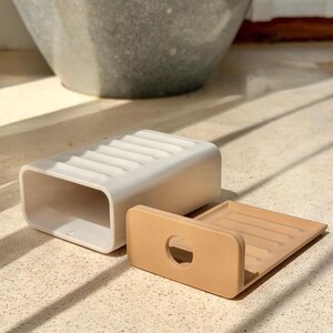 May include: A white rectangular soap dish with a ribbed surface and a hollow interior, accompanied by a separate tan-colored insert featuring a circular cutout and a matching ribbed design, both constructed from a smooth, solid material.