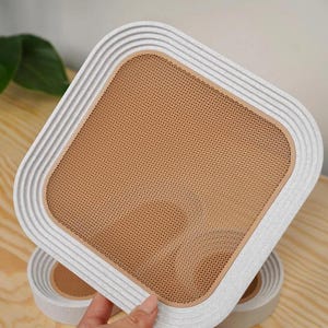 May include: A square, off-white cat scratcher with a textured, ridged border and a tan mesh center. The cat scratcher is held by a person's hand, with another one visible in the background. The design is modern and minimalist.