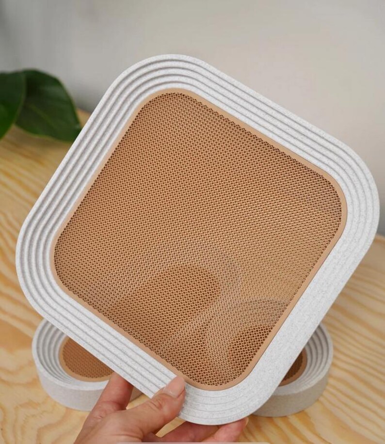 May include: A square, off-white cat scratcher with a textured, ridged border and a tan mesh center. The cat scratcher is held by a person's hand, with another one visible in the background. The design is modern and minimalist.