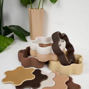 Puede incluir: Una colecci&oacute;n de macetas con forma de flor y posavasos a juego en varios tonos neutros. Las macetas est&aacute;n apiladas, con un jarr&oacute;n rectangular beige alto en el fondo. Los posavasos son en tonos marr&oacute;n, beige y blanco.