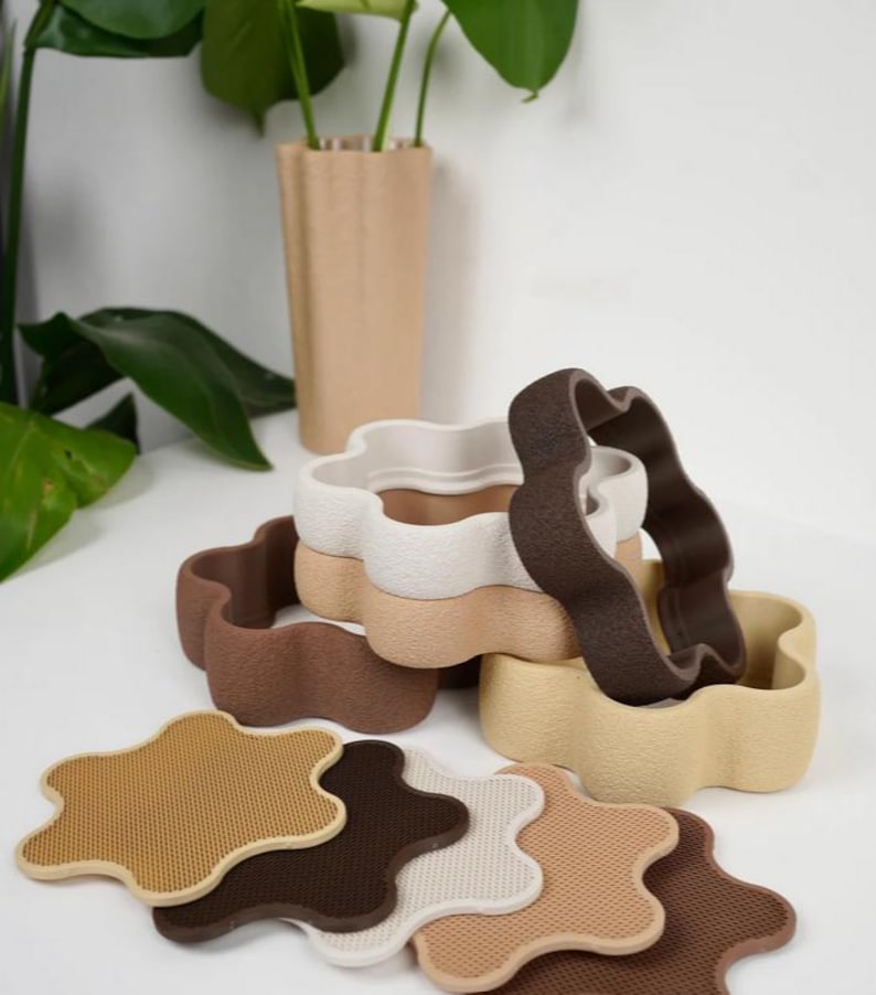 Puede incluir: Una colecci&oacute;n de macetas con forma de flor y posavasos a juego en varios tonos neutros. Las macetas est&aacute;n apiladas, con un jarr&oacute;n rectangular beige alto en el fondo. Los posavasos son en tonos marr&oacute;n, beige y blanco.