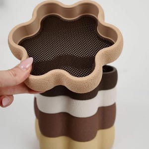 Puede incluir: Un recipiente multicapa con forma de flor, en tonos beige, blanco y marr&oacute;n. La capa superior, abierta, revela una malla. El dise&ntilde;o recuerda a un postre en capas, ofreciendo una est&eacute;tica &uacute;nica, con una altura aproximada de 15 cm.
