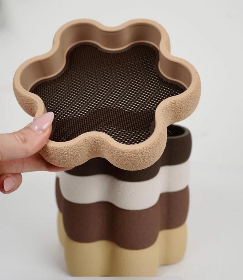 Puede incluir: Un recipiente multicapa con forma de flor, en tonos beige, blanco y marr&oacute;n. La capa superior, abierta, revela una malla. El dise&ntilde;o recuerda a un postre en capas, ofreciendo una est&eacute;tica &uacute;nica, con una altura aproximada de 15 cm.