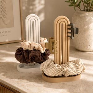 May include: Two arched hair accessory organizers, one white and one beige, each featuring a base and a vertical stand. The white organizer holds a beige ribbed scrunchie and a dark brown scrunchie. The beige organizer holds a waffle-textured scrunchie and two matte hair clips in olive and dark gray.