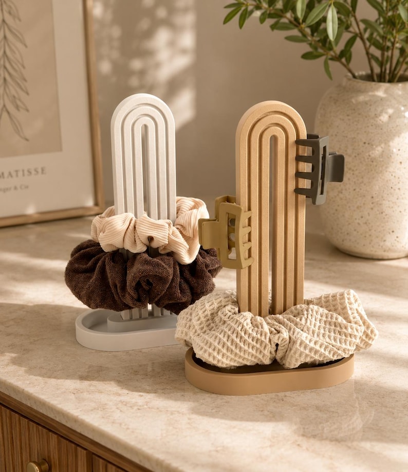 May include: Two arched hair accessory organizers, one white and one beige, each featuring a base and a vertical stand. The white organizer holds a beige ribbed scrunchie and a dark brown scrunchie. The beige organizer holds a waffle-textured scrunchie and two matte hair clips in olive and dark gray.