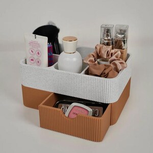 May include: A two-tone white and brown makeup organizer with a drawer. The organizer holds beauty products, including a white bottle, makeup brushes, and a tube of lotion. A drawer contains a makeup palette and a phone.