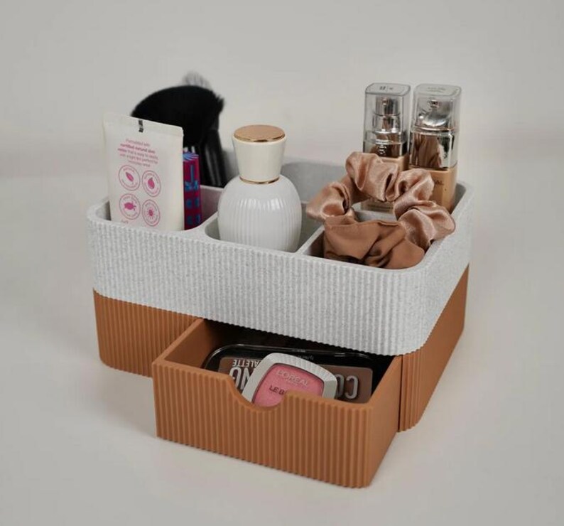 May include: A two-tone white and brown makeup organizer with a drawer. The organizer holds beauty products, including a white bottle, makeup brushes, and a tube of lotion. A drawer contains a makeup palette and a phone.