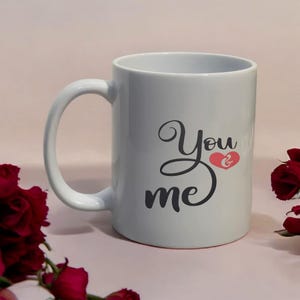 May include: White ceramic mug with a curved handle. The mug features the words "You & me" in a black script font, with a red heart symbol replacing the "&" symbol. The mug is surrounded by red roses.