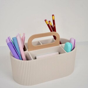 May include: Beige desk organizer with a handle, holding pens, pencils, and erasers. The organizer has multiple compartments and a ribbed exterior. The handle is a lighter shade of beige. The pencils are red with yellow accents, and the pens are various pastel colors.