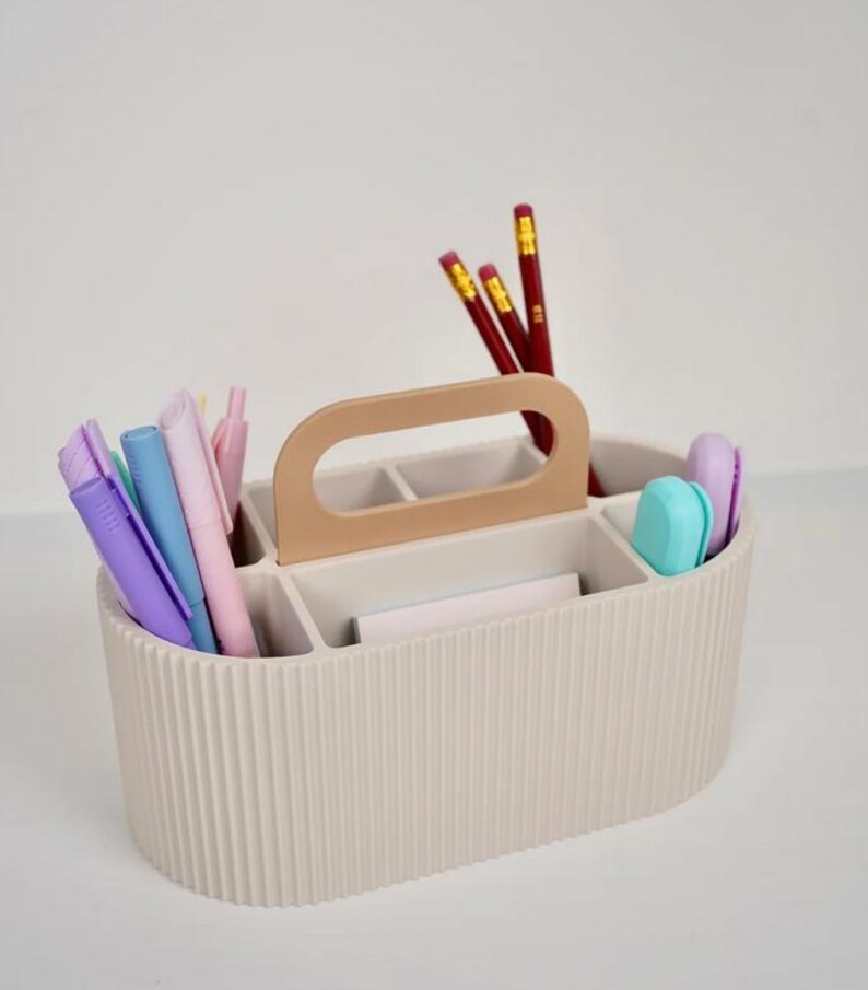 May include: Beige desk organizer with a handle, holding pens, pencils, and erasers. The organizer has multiple compartments and a ribbed exterior. The handle is a lighter shade of beige. The pencils are red with yellow accents, and the pens are various pastel colors.