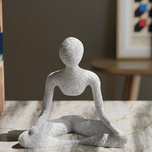 May include: A white, textured sculpture of a person in a seated yoga pose. The figure has a smooth, rounded head and arms extended to the sides. The sculpture is on a marble-patterned surface.