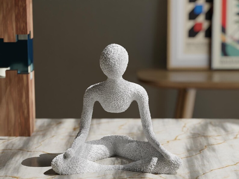 May include: A white, textured sculpture of a person in a seated yoga pose. The figure has a smooth, rounded head and arms extended to the sides. The sculpture is on a marble-patterned surface.