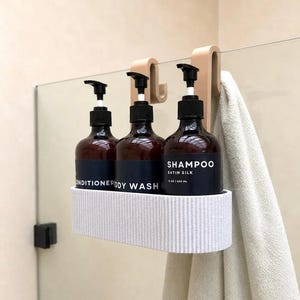 K&ouml;nnte beinhalten: Ein beiger Duschkorb zum Einh&auml;ngen an Glasw&auml;nden mit drei braunen Glasflaschen mit Pumpspender und schwarzen Etiketten mit der Aufschrift Conditioner, Body Wash und Shampoo Satin Silk 15 oz | 500 ml. Ein beiges Handtuch h&auml;ngt am gerippten Korb.