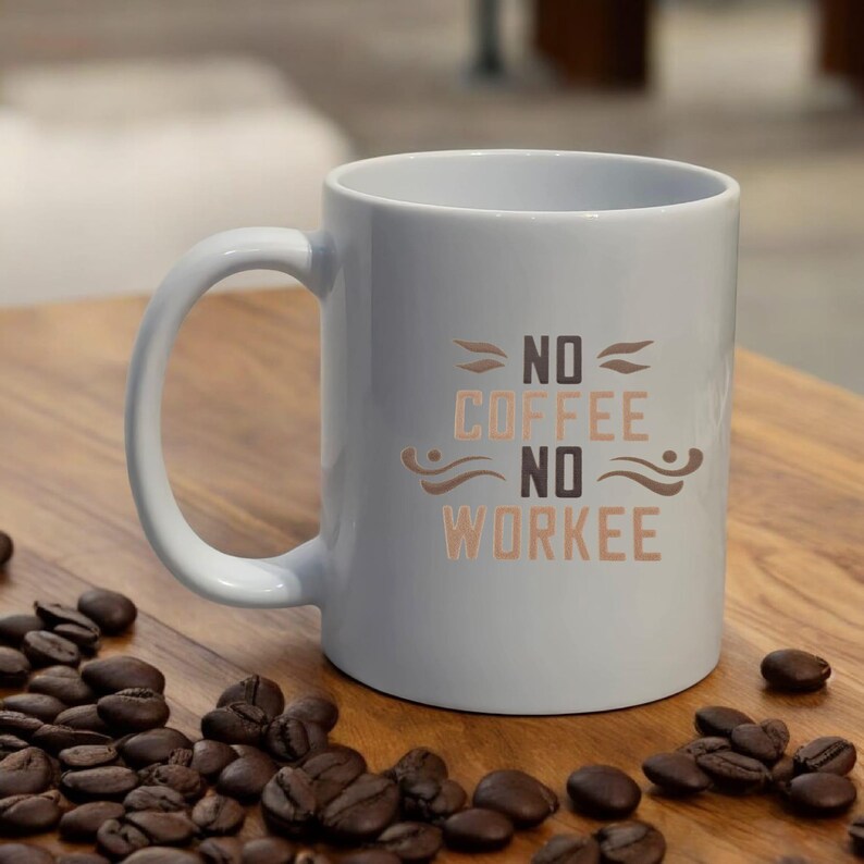 Puede incluir: Una taza de caf&eacute; de cer&aacute;mica blanca con el texto "NO COFFEE NO WORKEE" en letras marrones y beige. La taza tiene un asa curva y est&aacute; rodeada de granos de caf&eacute; sobre una superficie de madera.