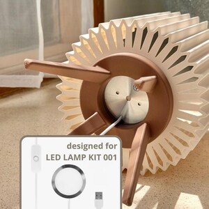 Puede incluir: Una pantalla de l&aacute;mpara circular plisada de color crema con una base marr&oacute;n de tres patas. Un gr&aacute;fico insertado muestra un cable de alimentaci&oacute;n blanco con un interruptor, un componente de luz LED circular y un conector USB, etiquetado "designed for LED LAMP KIT 001."
