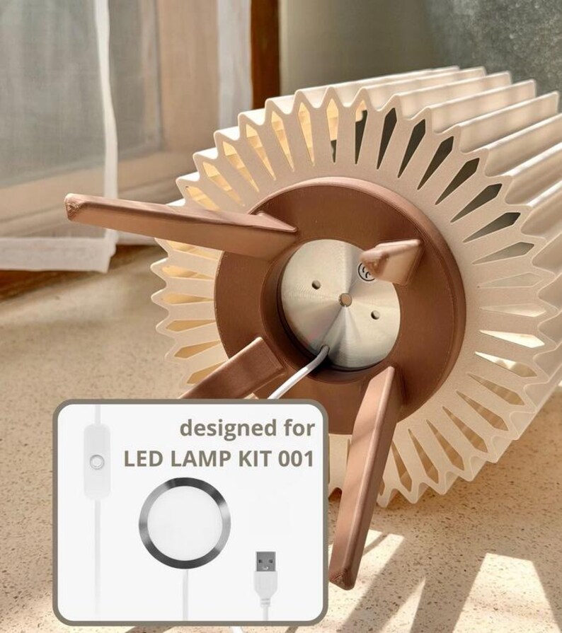 Puede incluir: Una pantalla de l&aacute;mpara circular plisada de color crema con una base marr&oacute;n de tres patas. Un gr&aacute;fico insertado muestra un cable de alimentaci&oacute;n blanco con un interruptor, un componente de luz LED circular y un conector USB, etiquetado "designed for LED LAMP KIT 001."