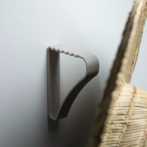 May include: A light gray, plastic hanger mounted on a white wall. The hanger has a ridged top and a curved design. A portion of a woven, light brown object is visible in the background.