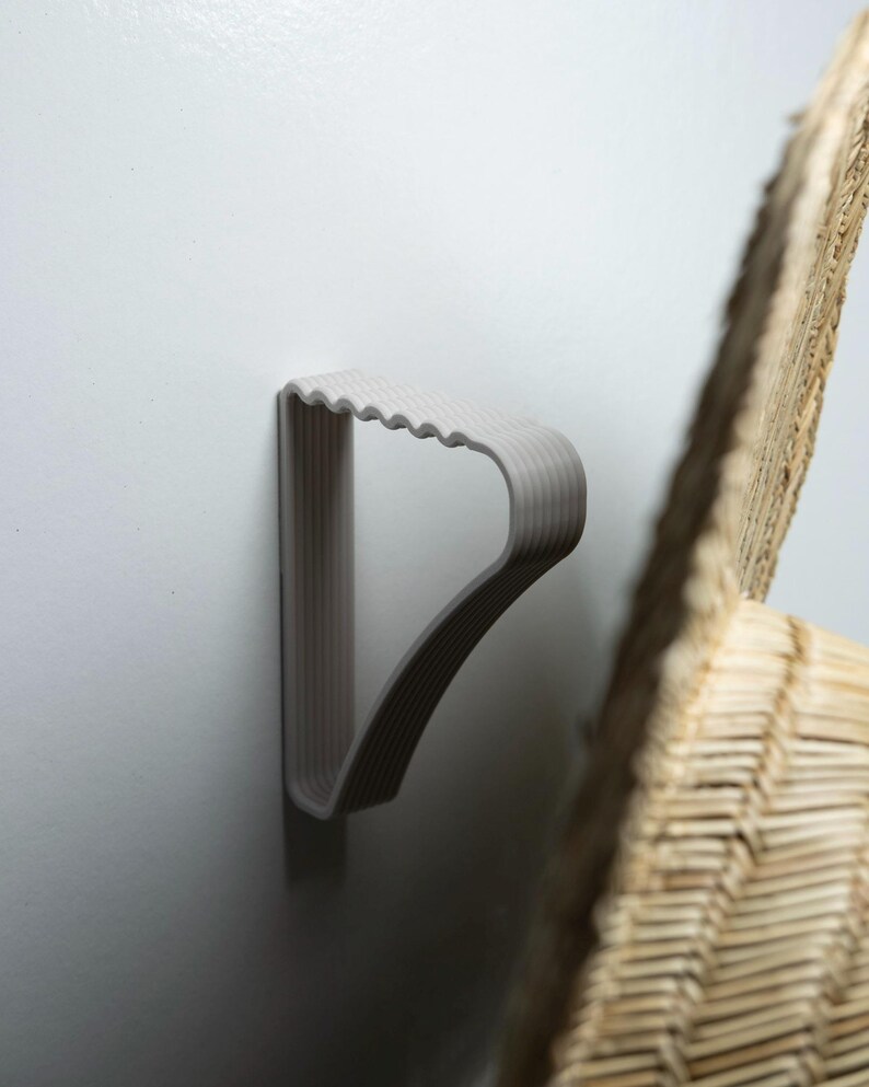 May include: A light gray, plastic hanger mounted on a white wall. The hanger has a ridged top and a curved design. A portion of a woven, light brown object is visible in the background.