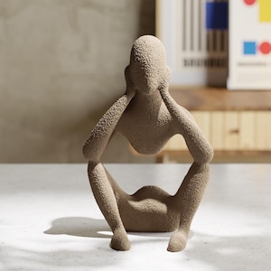 May include: A beige, textured sculpture of a seated figure with arms raised to the head. The figure has a rough, granular surface finish and a hollow torso area. The form is abstract, depicting a person in a crouched position with knees bent.