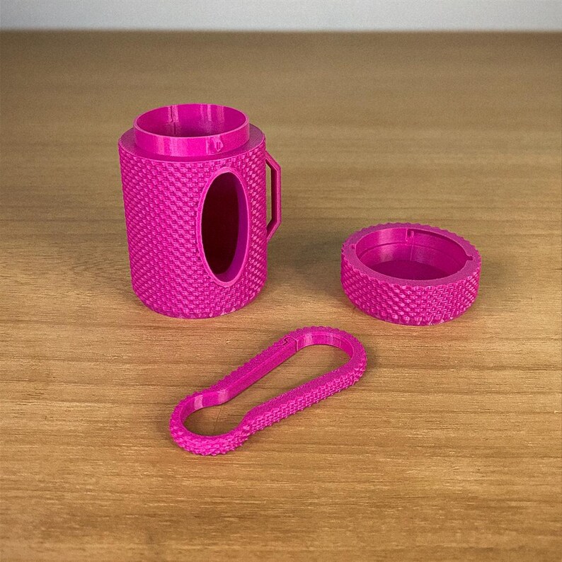 May include: A set of three magenta-colored 3D-printed accessories. The set includes a textured cylindrical container with a handle and an oval opening, a circular lid, and a carabiner-style clip. The items are arranged on a wooden surface.