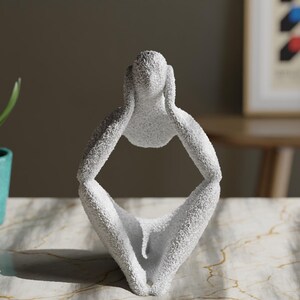 May include: A white, textured abstract sculpture of a seated figure with hands placed on the sides of the head. The figure has a rough, granular surface finish and a hollow center, depicting a person in a contemplative or resting pose.