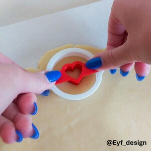 May include: A white circular cookie cutter with a red heart-shaped center insert being pressed into a flat sheet of light beige cookie dough. The tool features a circular outer frame and a central heart cutout, with the text @Eyf_design visible.