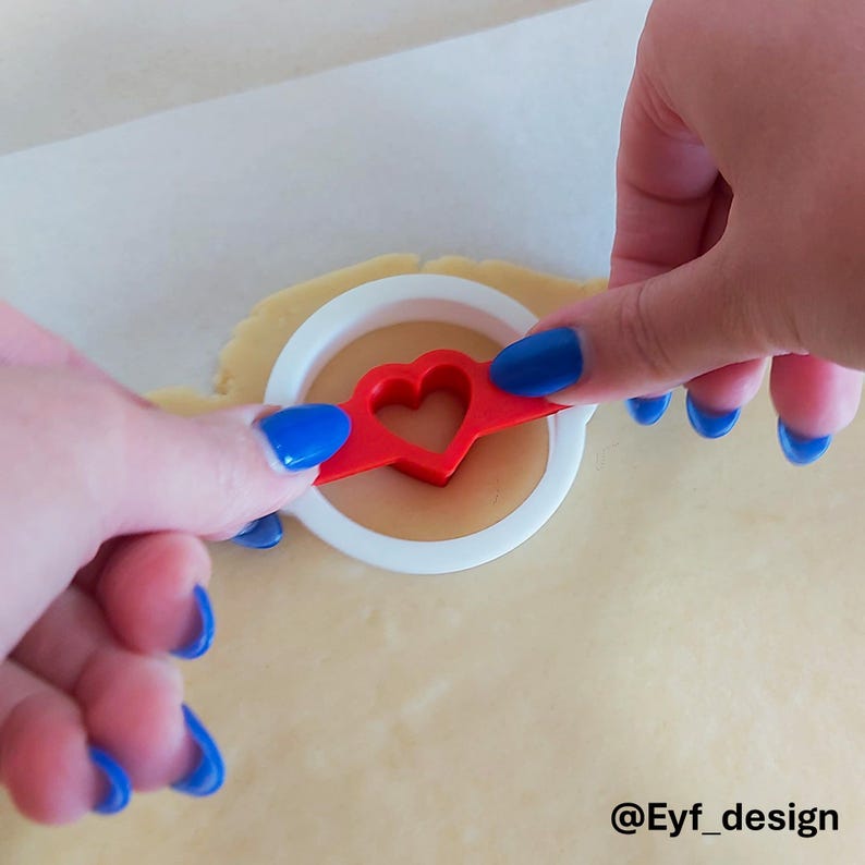 May include: A white circular cookie cutter with a red heart-shaped center insert being pressed into a flat sheet of light beige cookie dough. The tool features a circular outer frame and a central heart cutout, with the text @Eyf_design visible.