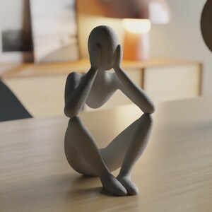 May include: A matte light gray sculpture of a seated human figure with a textured surface. The figure is depicted in a hunched position with both hands raised to cover the face, elbows resting on the knees, creating a contemplative posture.