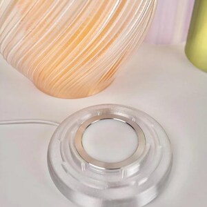May include: A clear, circular wireless charger with a silver ring around a white centre. A clear, swirled glass vase with an orange interior is in the background. The charger is connected to a white cord.