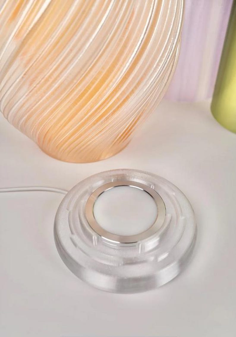 May include: A clear, circular wireless charger with a silver ring around a white centre. A clear, swirled glass vase with an orange interior is in the background. The charger is connected to a white cord.
