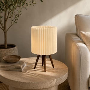 Puede incluir: Una l&aacute;mpara de mesa con una pantalla cil&iacute;ndrica de color blanco roto con pliegues verticales, sostenida por tres patas de madera de color marr&oacute;n oscuro. La l&aacute;mpara emite una luz c&aacute;lida y suave e incluye un cable de alimentaci&oacute;n blanco.