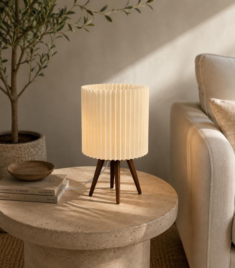 Puede incluir: Una l&aacute;mpara de mesa con una pantalla cil&iacute;ndrica de color blanco roto con pliegues verticales, sostenida por tres patas de madera de color marr&oacute;n oscuro. La l&aacute;mpara emite una luz c&aacute;lida y suave e incluye un cable de alimentaci&oacute;n blanco.