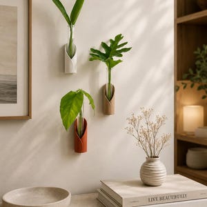 May include: Three wall-mounted plant propagation stations featuring cylindrical glass tubes held by curved white, tan, and terracotta-coloured holders. Each tube contains a single green plant cutting, displayed vertically on a plain wall surface.