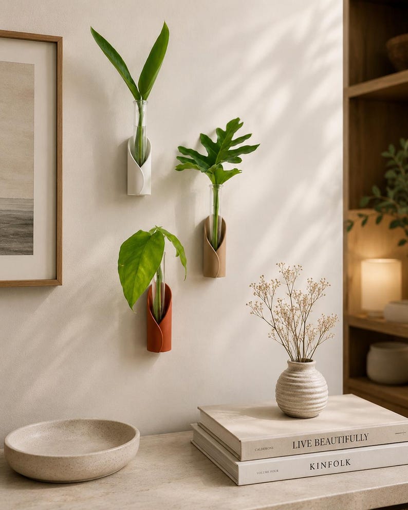 May include: Three wall-mounted plant propagation stations featuring cylindrical glass tubes held by curved white, tan, and terracotta-coloured holders. Each tube contains a single green plant cutting, displayed vertically on a plain wall surface.