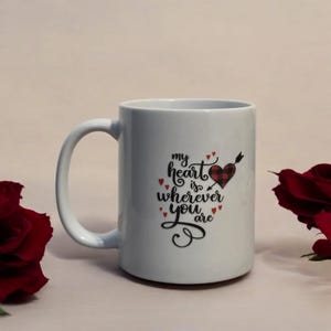May include: White ceramic mug with a large handle. The mug features the text "my heart is wherever you are" in black script, with a red plaid heart pierced by an arrow. Small red hearts accent the design.