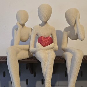 May include: Three abstract figures sit side-by-side on a shelf. The figures are a neutral beige color, with smooth, featureless faces. The center figure holds a red heart. Decorative shelf decor.