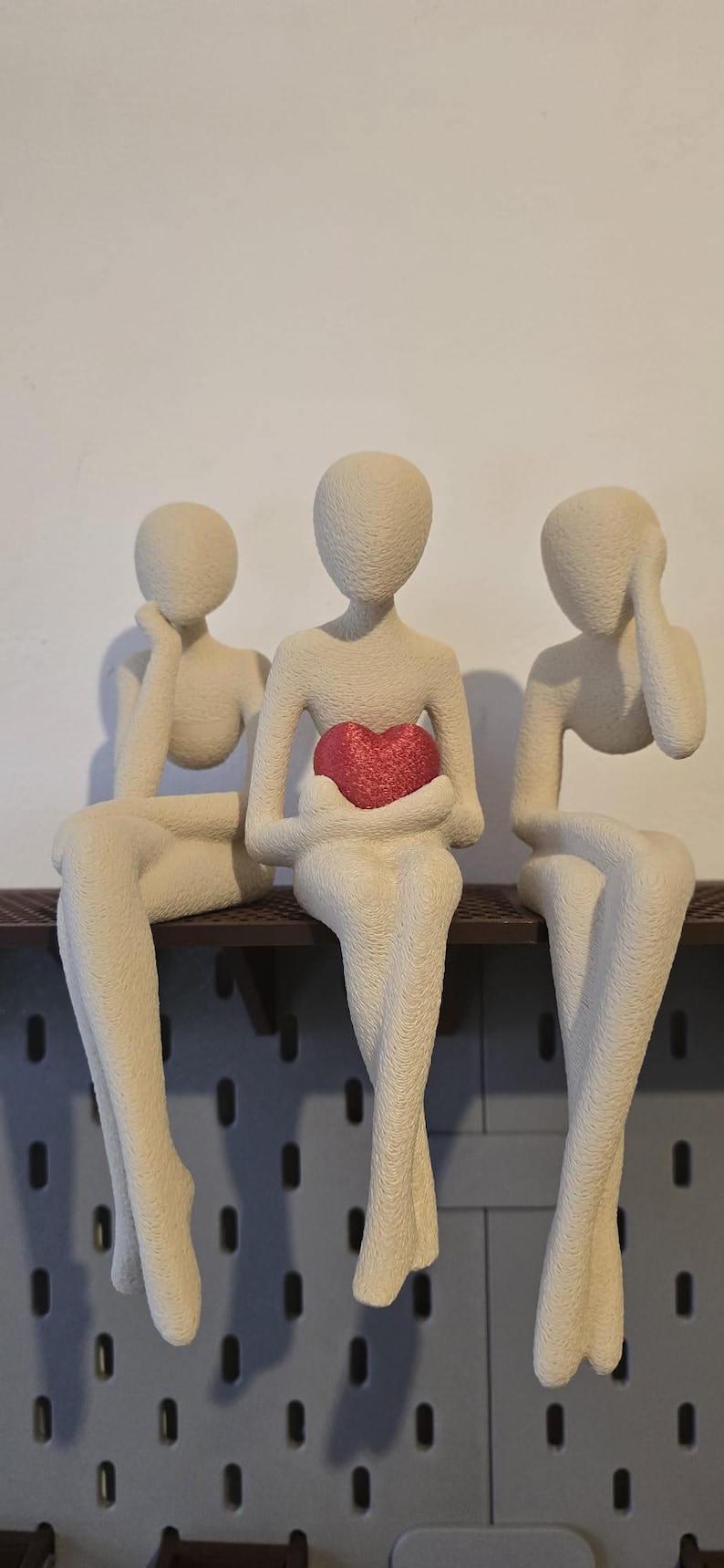 May include: Three abstract figures sit side-by-side on a shelf. The figures are a neutral beige color, with smooth, featureless faces. The center figure holds a red heart. Decorative shelf decor.
