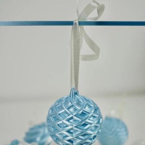 Pode incluir: Um ornamento esf&eacute;rico azul-claro com um padr&atilde;o de grade em relevo em forma de diamante. O ornamento est&aacute; suspenso por uma fita de cetim branca atada num la&ccedil;o. Outros ornamentos esf&eacute;ricos azuis e brancos s&atilde;o vis&iacute;veis no fundo desfocado.