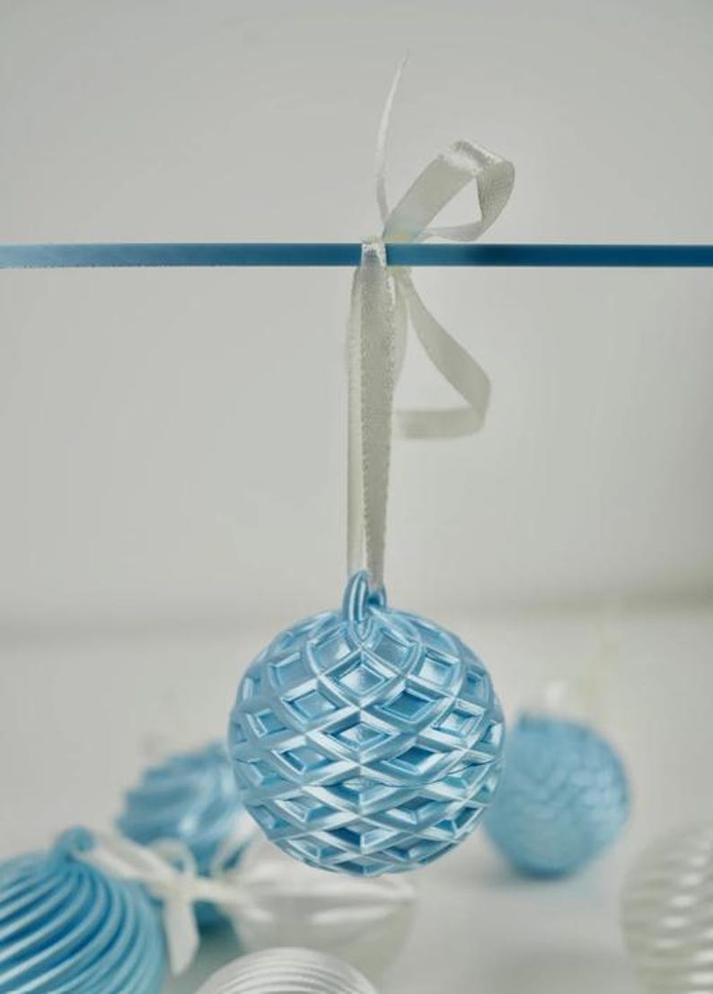 Pode incluir: Um ornamento esf&eacute;rico azul-claro com um padr&atilde;o de grade em relevo em forma de diamante. O ornamento est&aacute; suspenso por uma fita de cetim branca atada num la&ccedil;o. Outros ornamentos esf&eacute;ricos azuis e brancos s&atilde;o vis&iacute;veis no fundo desfocado.