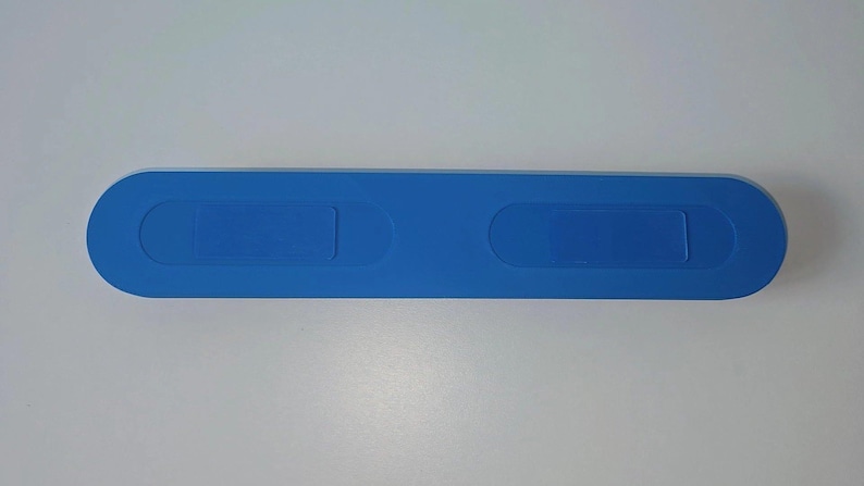 May include: A long, horizontal, blue plastic case with rounded ends. The top surface features two recessed, elongated oval sections positioned symmetrically along the center, providing a smooth, uniform appearance for this storage container.