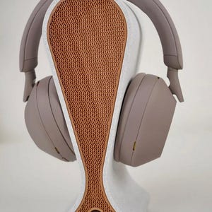 May include: A pair of taupe-colored headphones rests on a matching stand. The stand has a white base with a textured, orange-brown center. The headphones are designed to cradle the ears, and the stand is shaped to hold them securely.