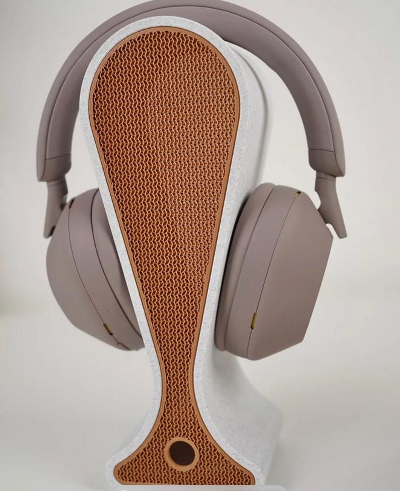 May include: A pair of taupe-colored headphones rests on a matching stand. The stand has a white base with a textured, orange-brown center. The headphones are designed to cradle the ears, and the stand is shaped to hold them securely.