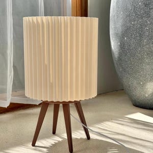 Puede incluir: Una l&aacute;mpara de mesa con una pantalla cil&iacute;ndrica plisada de color blanco roto, sostenida por tres patas de madera c&oacute;nicas de color marr&oacute;n oscuro. Un cable de alimentaci&oacute;n blanco sale de la base de la l&aacute;mpara, que se apoya en una superficie plana.