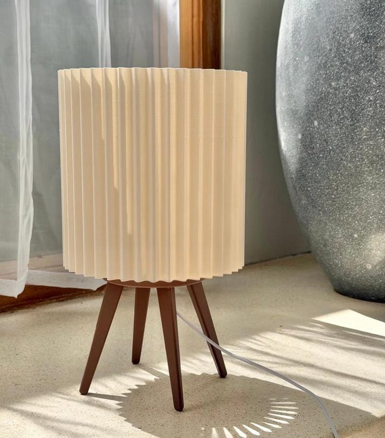 Puede incluir: Una l&aacute;mpara de mesa con una pantalla cil&iacute;ndrica plisada de color blanco roto, sostenida por tres patas de madera c&oacute;nicas de color marr&oacute;n oscuro. Un cable de alimentaci&oacute;n blanco sale de la base de la l&aacute;mpara, que se apoya en una superficie plana.