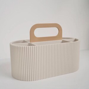 May include: A beige, ribbed, oval-shaped organizer with a light brown handle. The organizer has multiple compartments inside. The handle is a light brown color and is attached to the top of the organizer.
